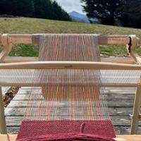 Woven cloth project with a patter including white, green, gold, and black yarn sitting on a woden table.